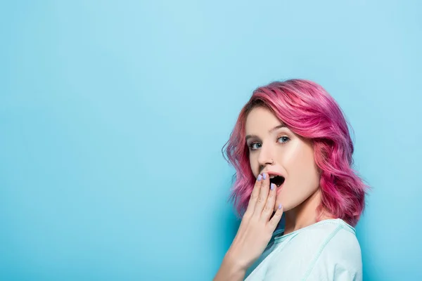 Chocado jovem mulher com cabelo rosa cobrindo boca no fundo azul — Fotografia de Stock