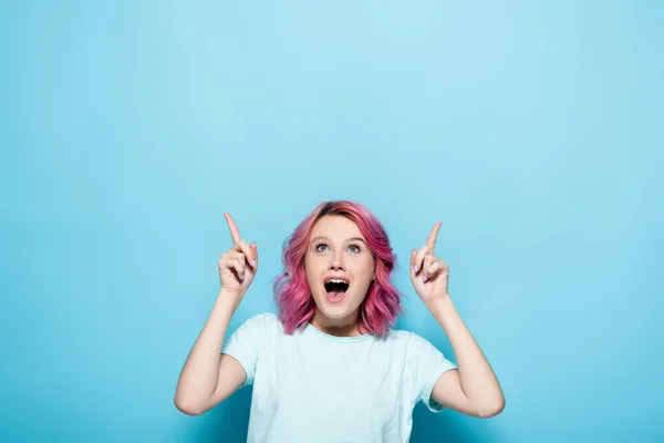 Choqué jeune femme avec les cheveux roses pointant vers le haut sur fond bleu — Photo de stock