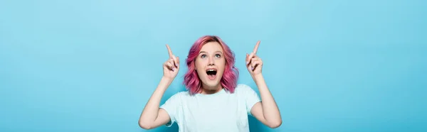 Chocado jovem mulher com cabelo rosa apontando para cima no fundo azul, tiro panorâmico — Fotografia de Stock