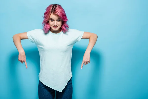 Jovem mulher com cabelo rosa apontando para baixo no fundo azul — Fotografia de Stock
