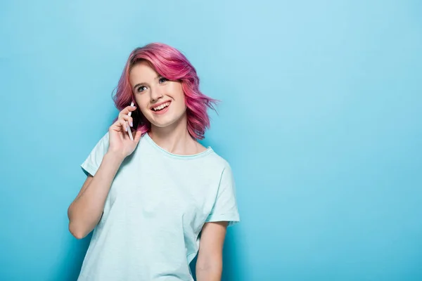 Mujer joven con el pelo rosa hablando en el teléfono inteligente y sonriendo sobre fondo azul - foto de stock