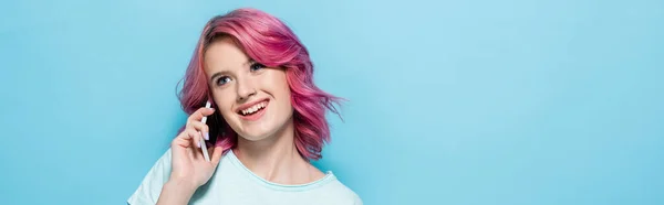 Mujer joven con el pelo rosa hablando en el teléfono inteligente y sonriendo sobre fondo azul, tiro panorámico - foto de stock