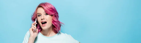 Young woman with pink hair talking on smartphone with open mouth isolated on blue, panoramic shot — Stock Photo