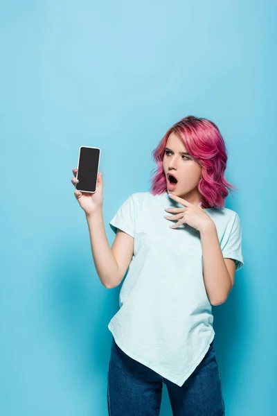 Шокированная молодая женщина с розовыми волосами держит смартфон с чистым экраном на синем фоне — стоковое фото