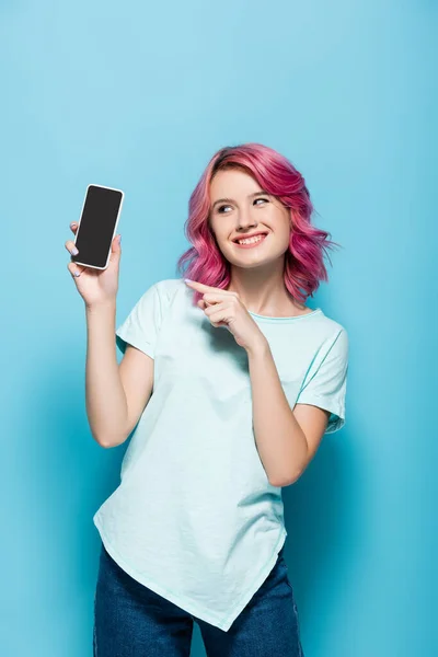 Mujer joven con el pelo rosa apuntando al teléfono inteligente con pantalla en blanco sobre fondo azul - foto de stock