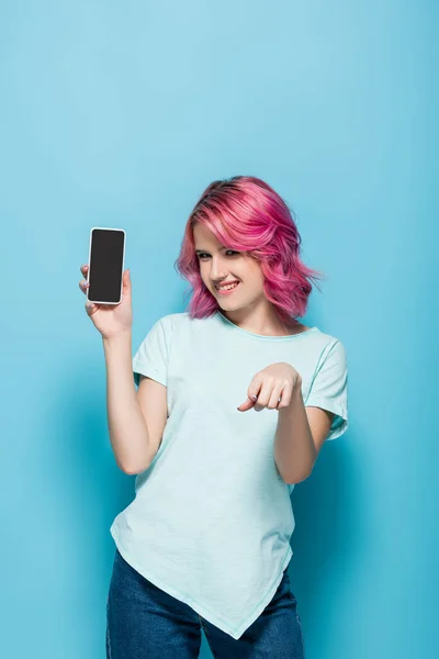 Mujer joven con pelo rosa sosteniendo teléfono inteligente con pantalla en blanco y sonriendo sobre fondo azul - foto de stock