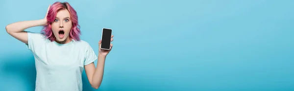 Shocked young woman with pink hair holding smartphone with blank screen on blue background, panoramic shot — Stock Photo