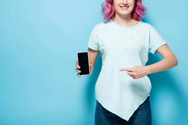 Vista recortada de la mujer joven con el pelo rosa apuntando al teléfono inteligente con pantalla en blanco sobre fondo azul, plano panorámico - foto de stock