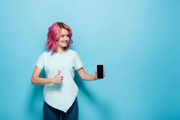 Junge Frau mit rosa Haaren hält Smartphone mit leerem Bildschirm und zeigt Daumen nach oben auf blauem Hintergrund — Stockfoto