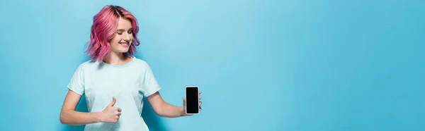 Mujer joven con pelo rosa sosteniendo teléfono inteligente con pantalla en blanco y mostrando el pulgar hacia arriba sobre fondo azul, plano panorámico - foto de stock