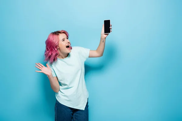 Збуджена молода жінка з рожевим волоссям тримає смартфон з порожнім екраном на синьому фоні — стокове фото