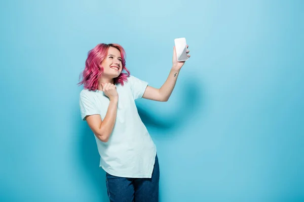 Junge Frau mit rosa Haaren hält Smartphone auf blauem Hintergrund — Stockfoto