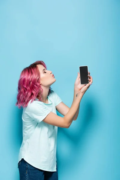 Молода жінка з рожевим волоссям цілує смартфон з порожнім екраном на синьому фоні — стокове фото