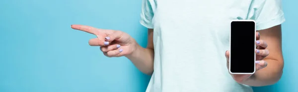 Cropped view of young woman holding smartphone with blank screen and pointing aside on blue background, panoramic shot — Stock Photo