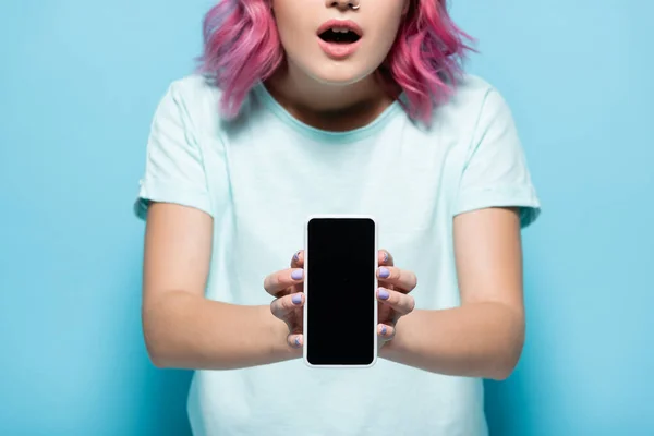 Vista recortada de la mujer joven conmocionada con el teléfono inteligente de celebración de pelo rosa con pantalla en blanco sobre fondo azul - foto de stock