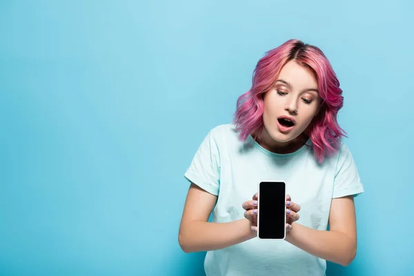 Schockierte junge Frau mit rosa Haaren hält Smartphone mit leerem Bildschirm auf blauem Hintergrund — Stockfoto