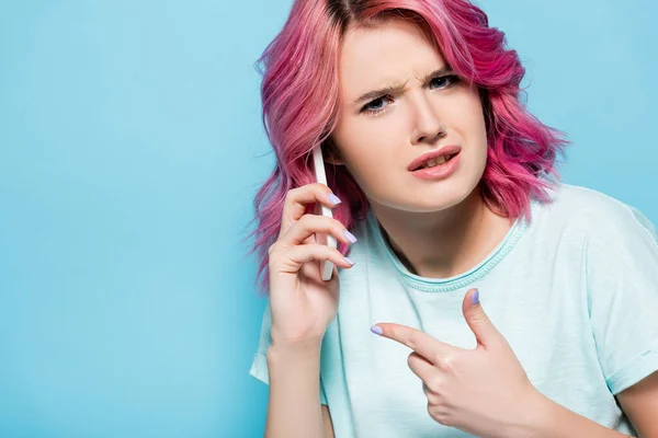 Mujer joven confundida con el pelo rosa apuntando a teléfono inteligente aislado en azul - foto de stock