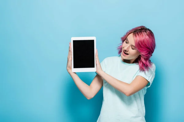 Überraschte junge Frau mit rosa Haaren präsentiert digitales Tablet mit leerem Bildschirm auf blauem Hintergrund — Stockfoto