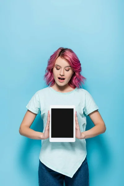 Aufgeregte junge Frau mit rosa Haaren präsentiert digitales Tablet mit leerem Bildschirm auf blauem Hintergrund — Stockfoto