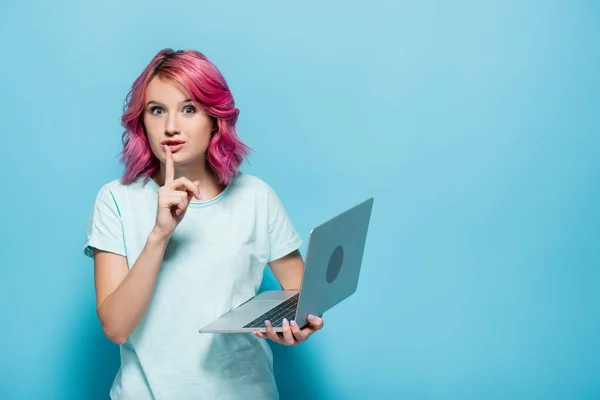 Mujer joven con el pelo rosa sosteniendo el ordenador portátil y mostrando el gesto shh sobre fondo azul - foto de stock