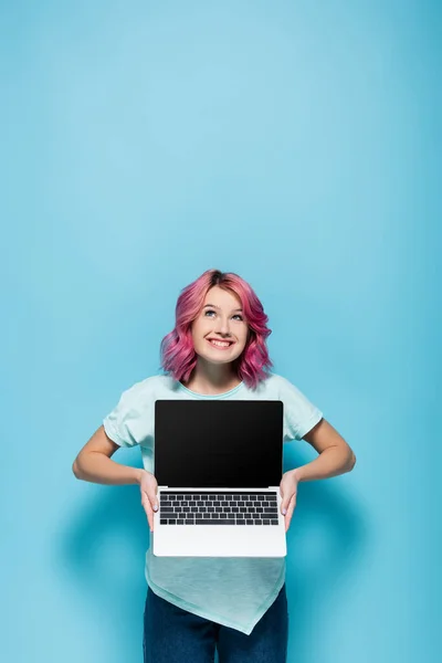 Young woman with pink hair showing laptop with blank screen and looking up on blue background — Stock Photo