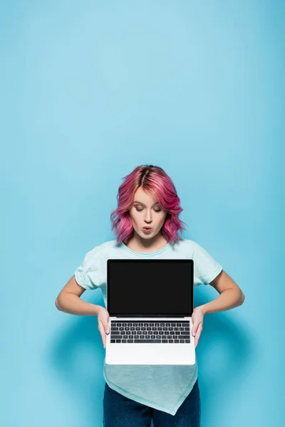 Choqué jeune femme avec les cheveux roses montrant ordinateur portable avec écran blanc sur fond bleu — Photo de stock