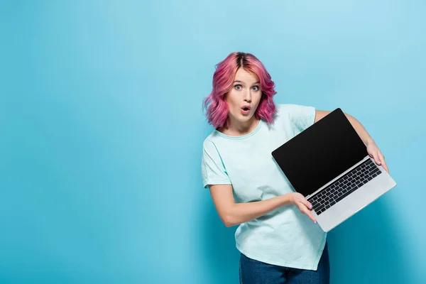 Шокированная молодая женщина с розовыми волосами показывает ноутбук с чистым экраном на синем фоне — стоковое фото