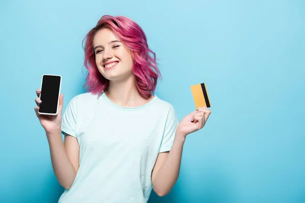 Jeune femme aux cheveux roses tenant carte de crédit et smartphone sur fond bleu — Photo de stock