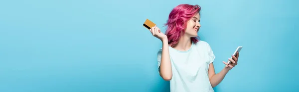 Молода жінка з рожевим волоссям тримає кредитну картку і смартфон на синьому фоні, панорамний знімок — стокове фото