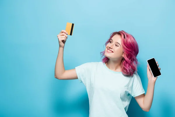 Giovane donna con capelli rosa in possesso di carta di credito e smartphone su sfondo blu — Foto stock