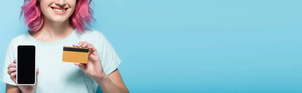 Vista ritagliata di giovane donna con capelli rosa in possesso di carta di credito e smartphone isolato su sfondo blu, colpo panoramico — Foto stock