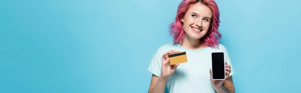 Young woman with pink hair holding credit card and smartphone on blue background, panoramic shot — Stock Photo