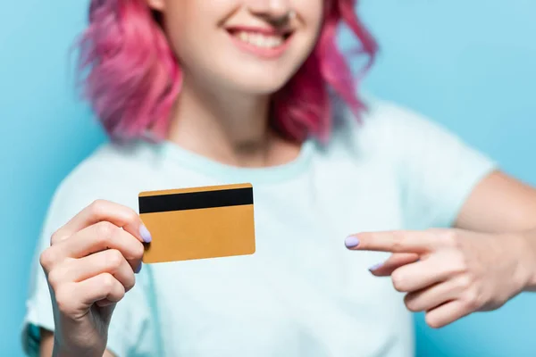 Enfoque selectivo de la mujer joven con el pelo rosa apuntando a la tarjeta de crédito sobre fondo azul - foto de stock