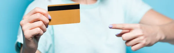 Foco seletivo da jovem apontando para o cartão de crédito em fundo azul, tiro panorâmico — Fotografia de Stock