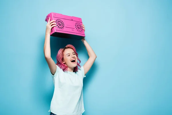 Mujer joven con pelo rosa sosteniendo grabadora de cinta vintage sobre fondo azul - foto de stock