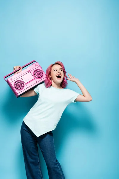 Excited young woman with pink hair holding vintage tape recorder on blue background — Stock Photo