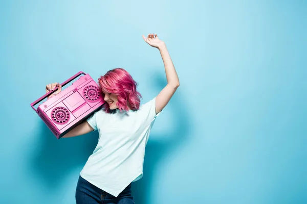 Mujer joven con pelo rosa sosteniendo grabadora de cinta vintage y bailando sobre fondo azul - foto de stock