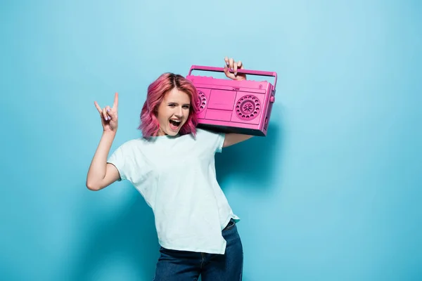 Mujer joven excitada con pelo rosa sosteniendo grabadora de cinta vintage y mostrando el signo de roca en el fondo azul - foto de stock