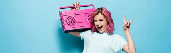 Mujer joven excitada con el pelo rosa sosteniendo grabadora de cinta vintage y mostrando el signo de roca en el fondo azul, tiro panorámico - foto de stock