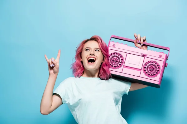 Mujer joven excitada con pelo rosa sosteniendo grabadora de cinta vintage y mostrando el signo de roca en el fondo azul - foto de stock