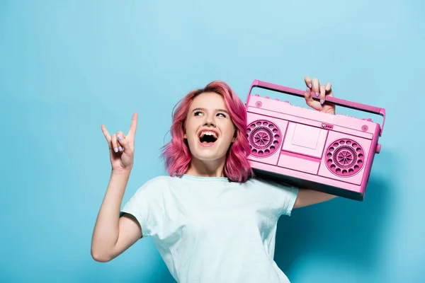 Eccitata giovane donna con i capelli rosa in possesso di registratore nastro vintage e mostrando segno di roccia su sfondo blu — Foto stock