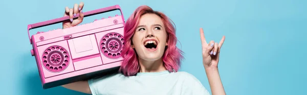 Mujer joven excitada con el pelo rosa sosteniendo grabadora de cinta vintage y mostrando el signo de roca en el fondo azul, tiro panorámico - foto de stock