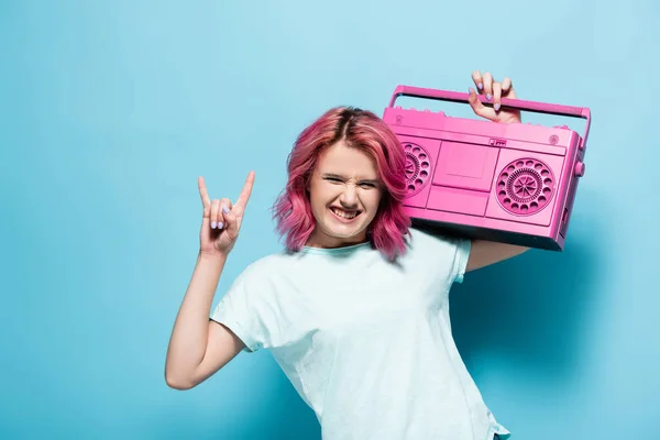 Junge Frau mit rosafarbenen Haaren hält Vintage-Tonbandgerät in der Hand und zeigt Rockzeichen auf blauem Hintergrund — Stockfoto