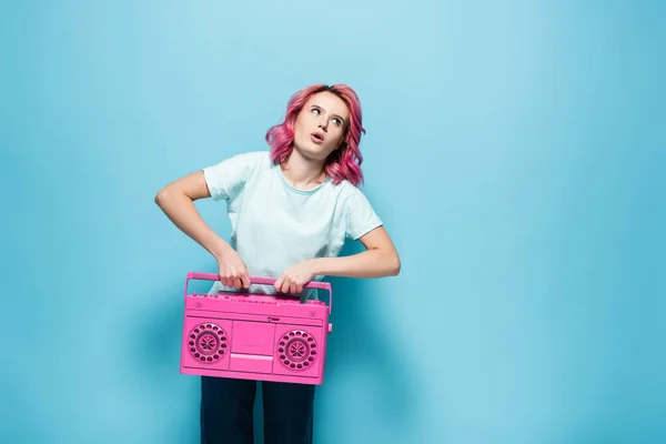 Young woman with pink hair holding heavy vintage tape recorder on blue background — Stock Photo