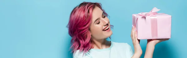 Young woman with pink hair holding gift box with bow and smiling on blue background, panoramic shot — Stock Photo