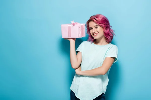 Молодая женщина с розовыми волосами держа подарочную коробку с луком и улыбаясь на синем фоне — стоковое фото