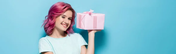 Young woman with pink hair holding gift box with bow and smiling on blue background, panoramic shot — Stock Photo