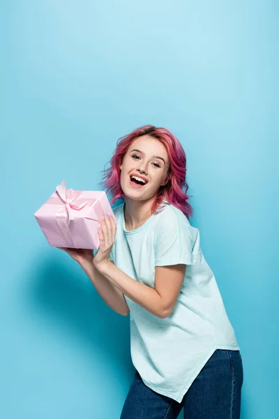 Aufgeregte junge Frau mit rosa Haaren hält Geschenkbox mit Schleife auf blauem Hintergrund — Stockfoto
