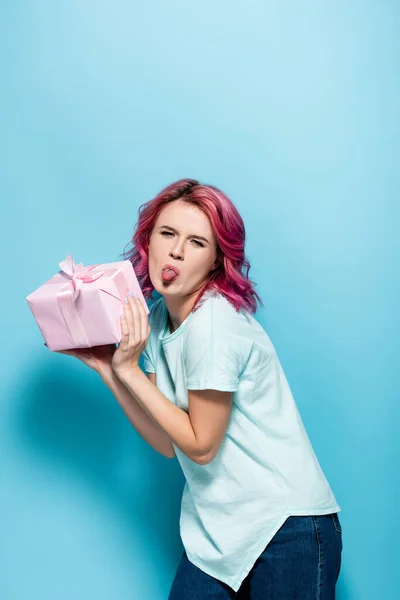 Young woman with pink hair holding gift box with bow and showing tongue on blue background — Stock Photo