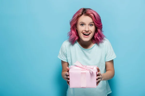 Excité jeune femme avec des cheveux roses tenant boîte cadeau avec arc sur fond bleu — Photo de stock